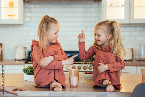 Two Scandinavian cute little sisters with ponytails sitting on kitchen table hugging knees, playing. Little swedish girls in pyjamas on breakfast time at home. Family domestic activities.