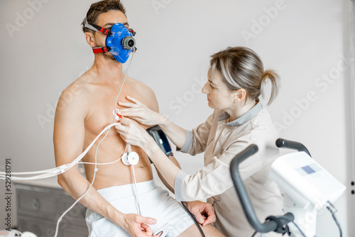 Nurse attaches electrodes to a man for a cardio endurance test during physical exercise on bike simulator, examining heart and vascular system. Man in breath mask on face
