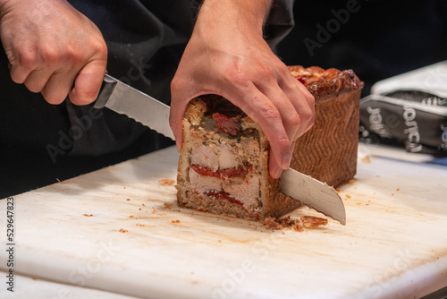 Male hand slicing with a big knife a pate en croute an appetizer meat pie wrapped in hot water crust pastry at a French market.