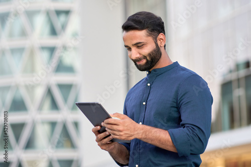 Smiling young adult indian business man professional, eastern businessman executive standing outdoors in urban city street checking data, holding using digital tablet online technology outside.