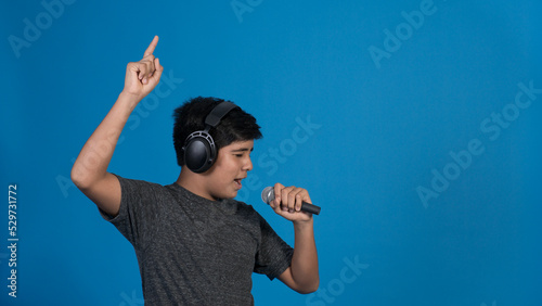 Retrato de estudio de un niño cantando con micrófono y audífonos mientras señala con dedo hacia arriba sobre fondo azul.