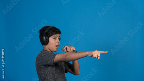 Retrato de estudio de un niño cantando con micrófono y audífonos mientras señala con dedo sobre fondo azul.