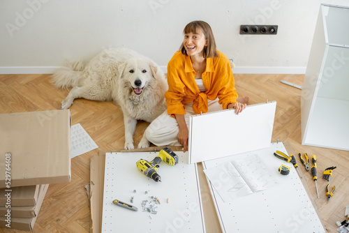 Portrait of a woman sits with her cute dog while assembling furniture at new apartment. DIY and house improvement concept