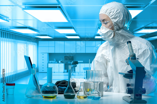 Bacteriological laboratories. Human in bacteriological protection suit. Study bacteria. Bacteriological woman is sitting at table. Girl in chemical protection suit in front of microscope and flasks
