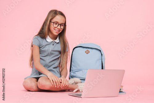 Happy caucasian young preteen schoolgirl teenage girl in uniform with bag using laptop e-learning remote studying education isolated in pink