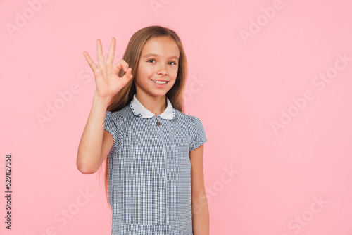 Young smart caucasian preteen teenager girl schoolgirl daughter in blue dress looking at camera showing okay gesture isolated in pink background