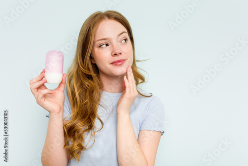 Young caucasian woman holding deodorant isolated on blue background looking sideways with doubtful and skeptical expression.