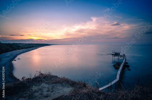 il tramonto sul trabocco di punta aderci, a vasto. il sole cala nell'entroterra abruzzese, dietro il gran sasso, e colora le nuvole ed il trabocco simbolo del parco. foto in lunga esposizione