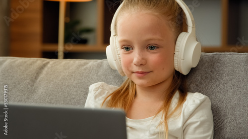 Funny little blonde girl child cute schoolgirl pretty beautiful kid young blogger wears headphones sit on sofa talking to webcam making online video call recording vlog having fun waving hand greeting
