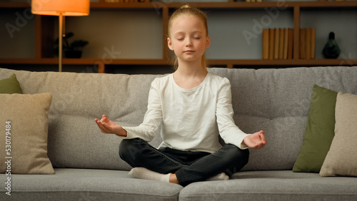 Little cute caucasian girl young healthy child blonde kid preschooler meditate sit on sofa at home calm mindful peaceful daughter do yoga breathing exercise relax alone with closed eyes in lotus pose