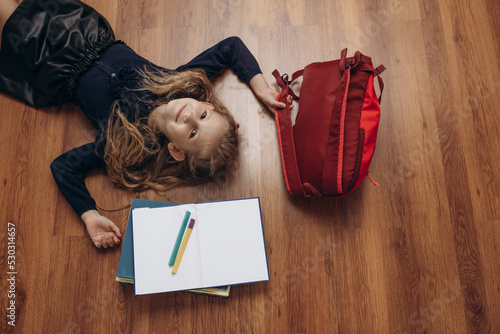 Beautiful girl child - schoolgirl with books, notebook, pens and backpack, portrait on the floor, top view
