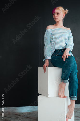 full body portrait of young woman with white and pink hair sitting on white cube on black background