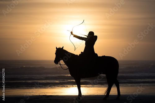 Mounted archer holds bow and arrow at sunrise on the beach.