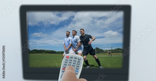 Composition of hand of sports fan holding remote control, with football match on tv screen