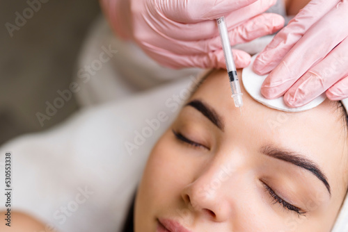 Close up of hands of young cosmetologist injecting botox in female face. She is standing and smiling. The woman is closed her eyes with relaxation