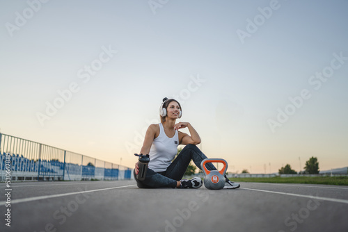 one caucasian woman female athlete rest hold supplement shaker kettlebell girya weight while sitting in stadium on track during training in summer evening copy space health and fitness concept