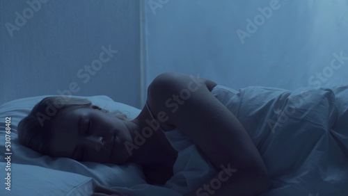 Young woman lying in the bed at night. Beautiful blond sleeping girl. Twilight in the bedroom, moonlight from the window. Health and rest concept.