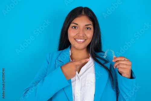 Young latin woman wearing blue blazer blue background holding an invisible aligner and pointing at it. Dental healthcare and confidence concept.