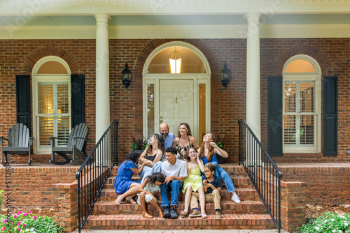 A large and blended and mixed happy family sitting on the front steps of their brick home in the suburbs