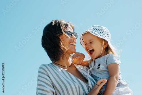 Positive family portrait laughing mom and daughter outdoors. Cheerful young beautiful mother holding laughing baby in her arms against blue sky