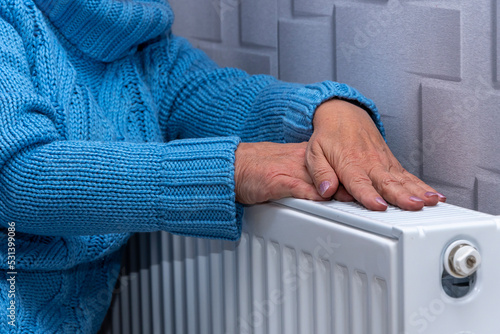 An elderly woman in a blue sweater warming her hands on a heating radiator. Concept: low room temperature, rising gas and heating prices, getting colder in the apartment.