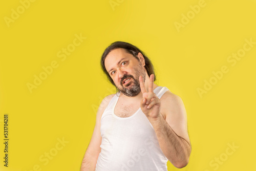 A disgruntled unshaven man in a white undershirt shows a two-finger peace gesture. Unemployed slovenly dressed man from Eastern Europe.