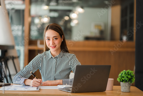 Smiling businesswoman working in office with money chart document and holding pen using laptop sitting on chair at studio in office accounting concept