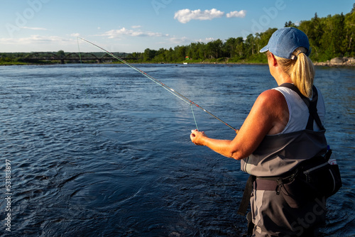 A middle age female stands in a large salmon river casting a fishing rod and holding line in her left hand. The blonde long haired lady is wearing a blue hat, white t-shirt, and waders fly-fishing. 