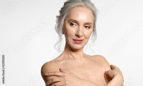 50s senior lady with perfect skin, closeup portrait. Elderly gray haired female touching naked shoulders on white background with copyspace. Aged beauty procedure and body care. Maturity and wellness