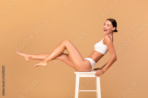 Overjoyed slim woman in white underwear sitting on white chair and smiling over beige studio background