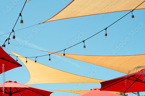 Multiple triangle shaped yellow nylon sunshades and awnings hanging over a patio deck. There are red colored canvas umbrellas hung with strings of clear patio light against a bright blue sunny sky.