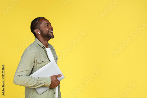 Laughing adult african american guy in wireless headphones with laptop looking up at empty space