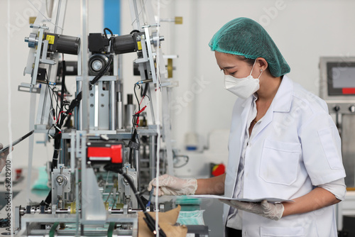 Worker woman in personal protective equipment or PPE inspecting quality of mask and medical face mask production line in factory, manufacturing industry and factory concept.
