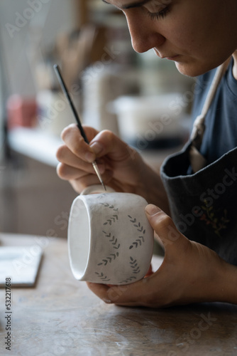 Hands of professional artisan hold white ceramic mug drawing pattern with dark paint using brush on blurred background. Woman enjoys working with handmade pottery craft in studio extreme closeup