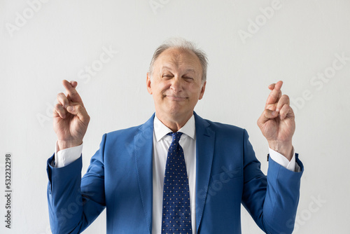 Portrait of dishonest mature businessman crossing fingers. Deceitful senior manager wearing formalwear looking at camera and smiling against white background. Liar concept