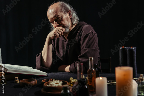 thoughtful alchemist reading magic cookbook near ingredients and candles isolated on black.