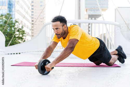 Handsome athletic african man training outdoors - Hispanic sportive man doing abs workout with ab roller wheel