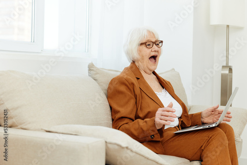 a happy, pleasantly shocked elderly woman is sitting on a sofa in a bright interior and looks at the camera with her mouth wide open, holding a laptop on her lap