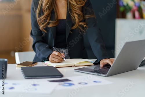 Close-up of a businesswoman sitting at a desk concentrating on fluently inspecting work using his laptop and several documents on the table at the office