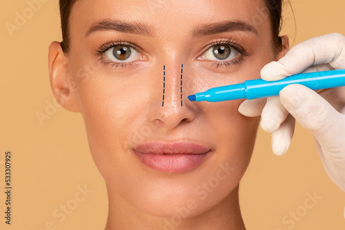 Rhinoplasty concept. Young woman with marking on her nose on beige studio background, closeup