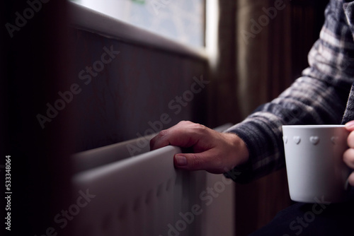 Close Up Of Mature Man Trying To Keep Warm By Radiator At Home During Cost Of Living Energy Crisis