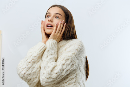 a beautiful, attractive, emotional woman stands on a light background in a white sweater and looks admiringly to the side touching her face with her hands