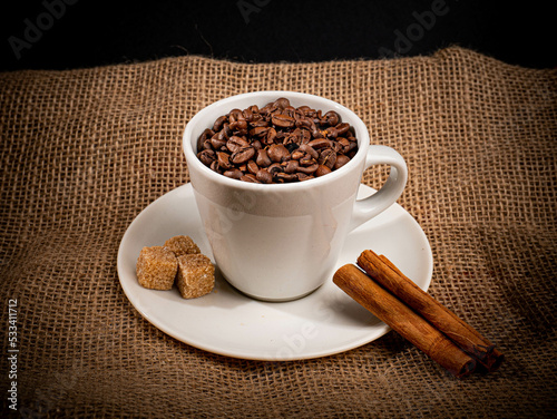 Ziarna kawy w białej filiżance na białym talerzyku z brązowym cukrem oraz cynamonem Postawione na worku jutowym po kawie