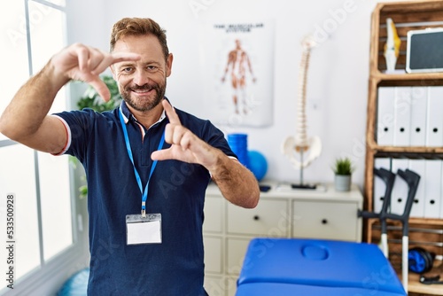 Middle age physiotherapist man working at pain recovery clinic smiling making frame with hands and fingers with happy face. creativity and photography concept.