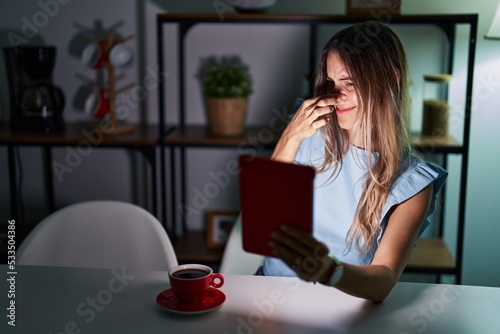 Young hispanic woman using touchpad sitting on the table at night smelling something stinky and disgusting, intolerable smell, holding breath with fingers on nose. bad smell