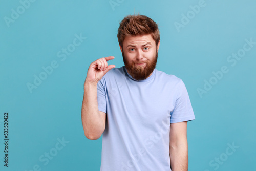 Portrait of disappointed bearded man showing a little bit gesture, dissatisfied with low rating, measuring scale, frowning face and looking at camera. Indoor studio shot isolated on blue background.