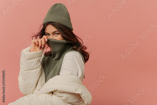 Cheerful young caucasian girl looking into cameras covers her face with collar of sweater on pink background. Brunette with wavy hair wears grey hat, sweater and white jacket. 