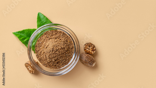 Ground nutmeg powder in a glass bowl and seeds over beige background. Grated muscat powder for spice and seasoning concept. Myristica fragrans tree fruit for herbal medicine. Copy space.