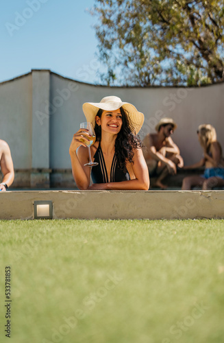 vertical photo of a smiling young woman wearing a pamela and leaning on the edge of a pool