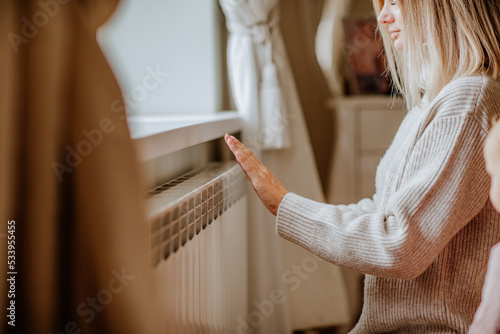 Young woman in long winter beige sweater is posing at home near the radiator